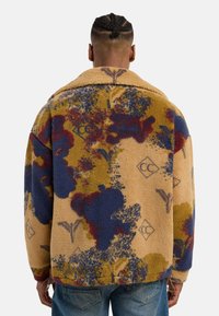 Sædvanlig beige fleecejakke med flerfarvede abstrakte mønstre i blå, maroon og gul. Har en stor krave og en afslappet pasform.