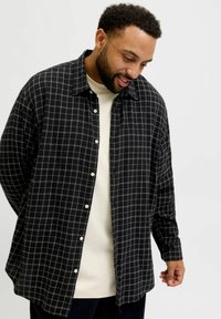 Mees vaatab alla, seljas must ruuduline särk valge T-särgi üle, taustal lihtne hele taust.