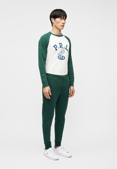 Νεαρός άνδρας που στέκεται, φορώντας λευκό και πράσινο μακρυμάνικο πουκάμισο με το κείμενο «P.R.L », σκούρα πράσινα παντελόνια jogger και λευκά αθλητικά παπούτσια.