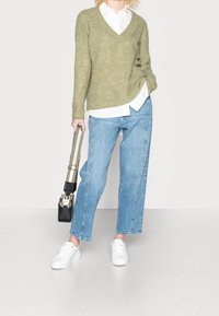 Grüner Strickpullover mit V-Ausschnitt, darunter ein weißes Hemd mit Kragen, hellblaue Jeans mit weitem Bein, weiße Turnschuhe und eine kleine schwarze Umhängetasche.