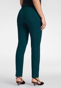 Grüne, figurbetonte Hose mit glatter Textur, mit mittelhohem Bund und Seitennähten. Kombiniert mit braunen, schnürenden High Heels.