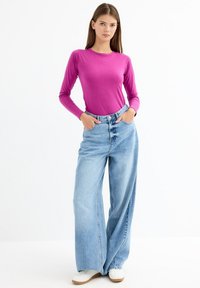 Top de manga larga ajustado en rosa brillante, combinado con jeans de pierna ancha en azul claro. Modelo de pie con las manos en los bolsillos, usando zapatillas blancas.