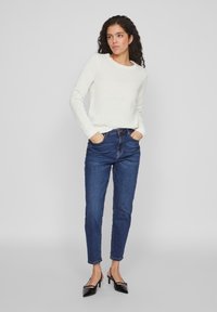 Pulover alb din tricot cu guler rotund, asortat cu blugi închiși de culoare albastră cu talie înaltă. Pantofii cu vârf ascuțit negri completează ținuta.
