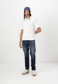 Vit kortärmad polo med ribbad textur, kombinerad med blå jeans och off-white sneakers. Modellen bär en blå mössa.