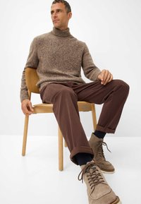 Brauner Strickrollkragenpullover, dunkelbraune Hose, graue Wildlederschuhe mit Schnürsenkeln, sitzt auf einem hellen Holzstuhl vor einem schlichten Hintergrund.