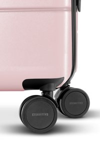 Valigia rigida di colore rosa chiaro con una finitura texture. Presenta quattro ruote nere e dettagli in plastica, etichettata "ETERNITIV3."