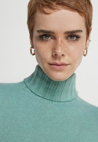 Turkos polotröja med ribbad textur. Närbild på halsringningen och ansiktet, som har fräknar och stora örhängen.