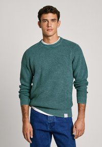 Zeleno pleteno pulover s teksturiranim vzorcem, okroglo izrezan, z rebrastimi manšetami. Nošen čez belo srajco, v kombinaciji z modrimi jeans hlačami.