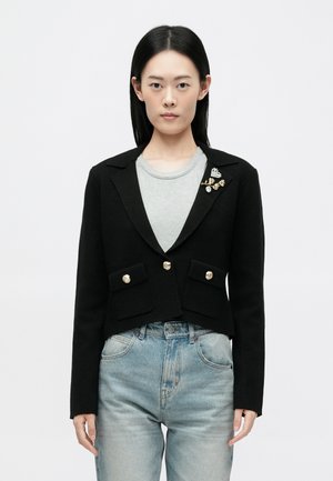 Fekete cropped blézer arany gombokkal és két elülső zsebből. Díszes brosst tartalmaz gyöngyökkel és arany részletekkel. Szürke felsőre van felvéve.