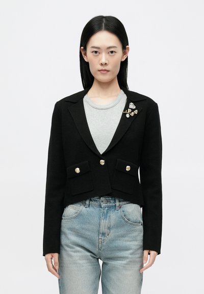 Zwarte cropped blazer met gouden knopen en twee voorzakken. Bevat een decoratieve broche met parels en gouden accenten. Gedragen over een grijze top.