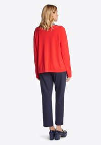 Roter Strickpullover mit rundem Halsausschnitt, kombiniert mit navyfarbener Hose. Das Model trägt navyfarbene Plateau-Schuhe und wird von hinten gezeigt.