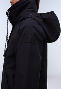 Persona con chaqueta negra con capucha, cuello alto y cordón, vista de lado que muestra la parche del logo en la manga.