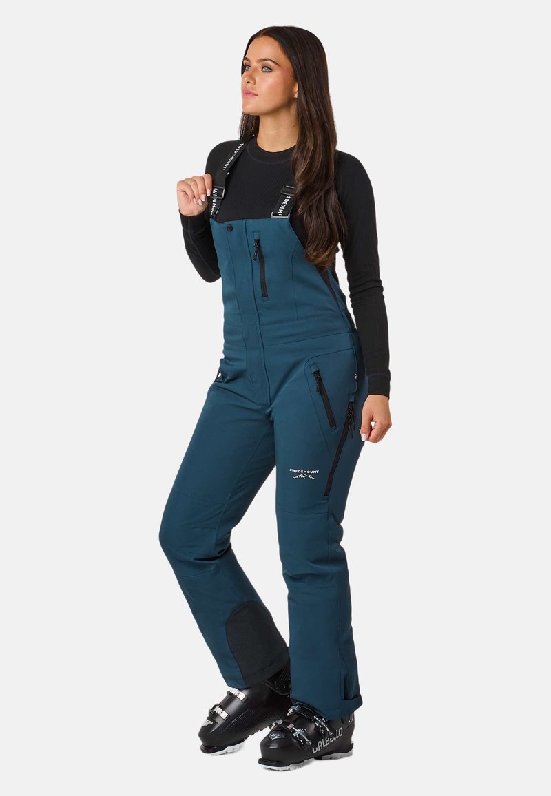 Türkise Skioveralls mit verstellbaren Trägern, Frontreißverschluss und Seitentaschen. Verstärkte Knieflicken; wasserabweisender Stoff. Eng anliegende Passform.