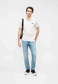 Άσπρο πόλο μπλουζάκι με μαύρο λογότυπο, ανοιχτό μπλε τζιν και μαύρα αθλητικά παπούτσια. Το μοντέλο κρατά μια μαύρη τσάντα ώμου, στέκεται μπροστά σε μια απλή φόντο.