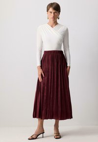 Jupe midi plissée bordeaux avec une taille élastique, assortie à un haut ajusté à manches longues blanc. Le mannequin porte des talons noirs à lanières.