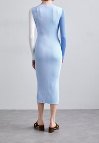 Žena stojí proti šedé zdi a má na sobě pletené midi šaty v barevném provedení s bílými a modrými dlouhými rukávy a hnědými sandály na podpatku.