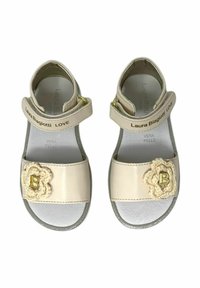 Sandali in pelle beige con cinturini regolabili, accessori in oro e un dettaglio di fiore all'uncinetto che presenta una "B" dorata sulla cinghia del dito.