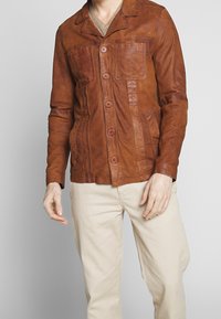 Brun läderjacka med kragdesign, knappframsida och två bröstfickor. Ytan verkar slät med en något sliten finish.