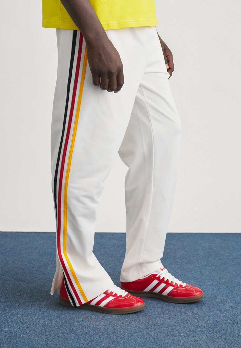 Άτομο που φοράει λευκό παντελόνι με κίτρινες, κόκκινες και μαύρες πλε υρικές ρίγες και κόκκινα αθλητικά παπούτσια με λευκές ρίγες σε μπλε χαλί.