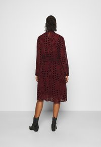 Röd och svart mönstrad klänning med långa ärmar och åtsittande midja; medellång, svängande tyg med subtil struktur; bärs med svarta ankelstövlar.