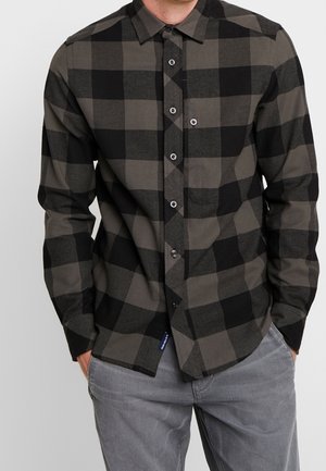 Homme portant une chemise boutonnée à manches longues avec un grand motif à carreaux noirs et gris foncé et un pantalon gris clair, mains dans les poches.
