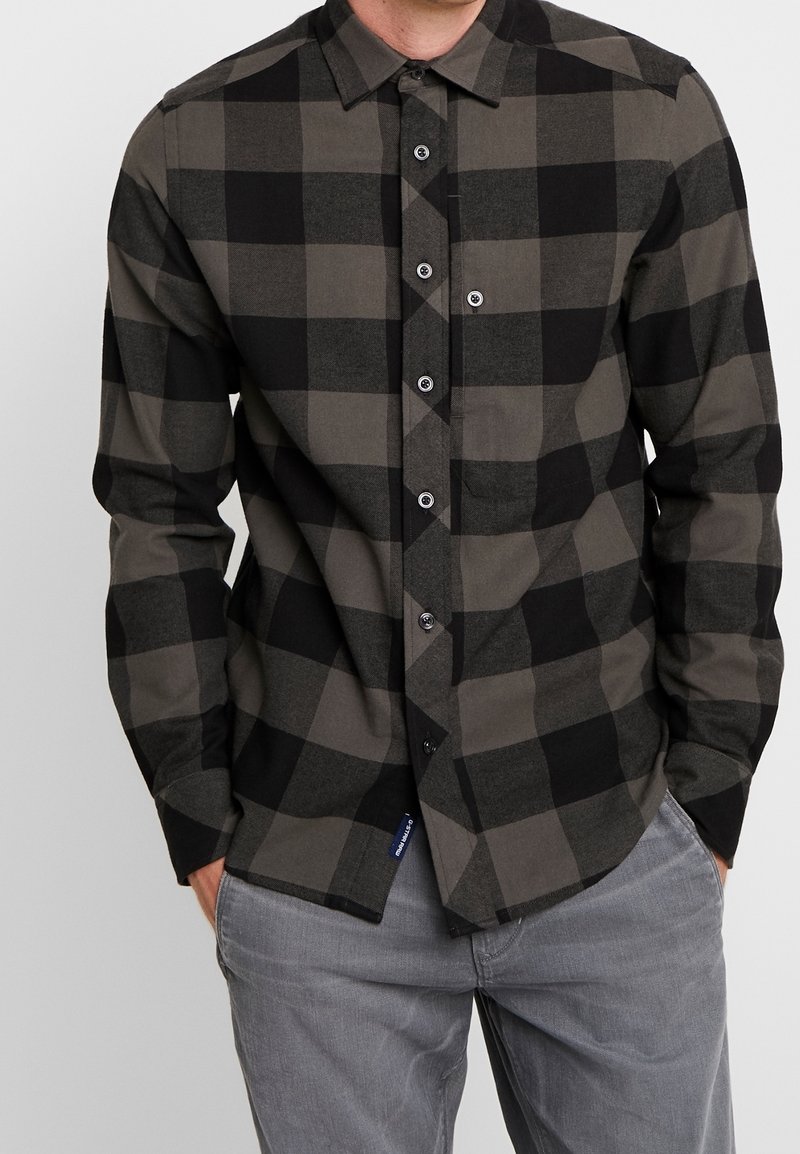 Homme portant une chemise boutonnée à manches longues avec un grand motif à carreaux noirs et gris foncé et un pantalon gris clair, mains dans les poches.