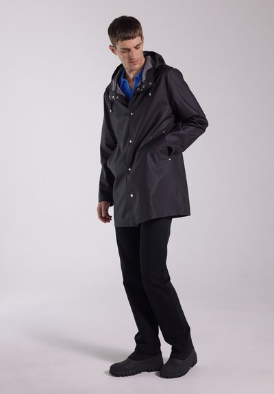 Black waterproof raincoat with a gray hood, snap button closures, side pockets, and a relaxed fit. Model wearing dark pants and shoes.
