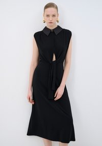 Robe noire avec un col, design sans manches, ouverture en clé à l'avant et détail de taille noué. Tissu lisse et coupe jusqu'à la cheville.