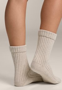 Rżebrowane kremowe skarpetki z podwiniętym mankietem, prezentujące teksturowany wzór dzianiny. Posiadają ciasne dopasowanie wokół kostki i w połowie łydki.