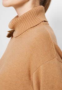 Personne portant un pull à col roulé côtelé de couleur camel avec le col replié, montrant un profil latéral de la tête aux épaules.