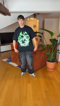 Juodas oversize stiliaus marškinėlis su žaliais grafikos kaukolės atvaizdais ir šešėliais. Laidūs tamsūs cargo stiliaus kelnės ir pilkos sportbačiai. Pastatytas vidaus erdvėje, kur yra augalų.