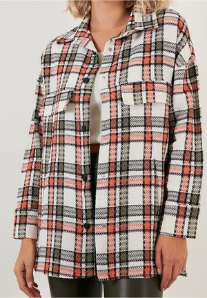 Femme portant une veste à carreaux boutonnée avec des rayures orange, noire et vert olive, par-dessus un haut blanc et un pantalon noir.