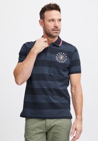 Polo tričko v námorníckej modrej so pruhmi, s golierom, ktorý má červené a biele akcenty, vyšitým logom na hrudi a krátkymi rukávmi.