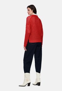 Roter Häkelpullover mit lockerem Sitz, gerippten Bündchen und Saum, kombiniert mit dunkler Jeanshose und weißen Stiefeln mit Absatz.