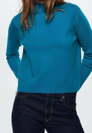 Pull en maille côtelée turquoise avec un col haut et des manches longues, associé à un jean en denim foncé. Texture lisse, coupe décontractée.