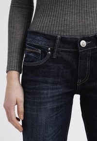 Jeans di denim scuro con design aderente, caratterizzati da leggere sfumature, tasche tradizionali e chiusura con bottone metallico. Top grigio a coste con trama.