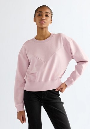Jeune femme aux cheveux tressés portant un sweat-shirt rose clair et un pantalon noir, posant avec une main sur la hanche devant un fond blanc.