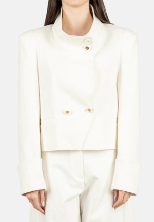 Donna che indossa un blazer bianco su misura con bottoni dorati e pantaloni ampi abbinati, mostrata dal collo ai fianchi, in piedi con le braccia rilassate.