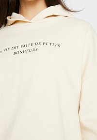 Krämfärgad hoodie med svart text som säger: "La vie est faite de petits bonheurs." Tyget ser mjukt ut med en avslappnad passform.