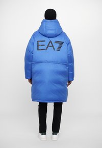 Garšs zils pūkainais mētelis ar kapuci, aizmugurē ar melnu 'EA7' logotipu. Izgatavots no gludas, spīdīgas materiāla, līdz ceļiem.