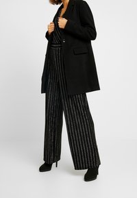 Femme portant un long manteau noir par-dessus une combinaison à rayures verticales noires et argentées, avec des bottines à talons noirs.