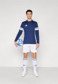 Marineblauw sportshirt met lange mouwen en witte strepen, gecombineerd met witte shorts en kniehoge sokken met blauwe accenten, terwijl hij een voetbal vasthoudt.