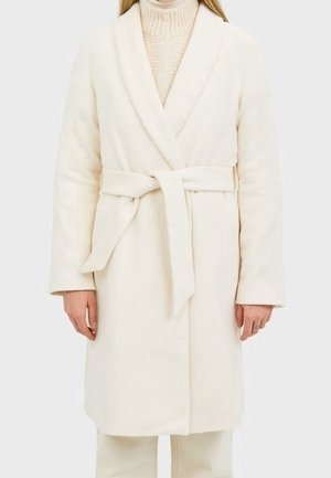 Femme portant un manteau en laine crème ceinturé sur un pull à col roulé clair, debout devant un fond uni.