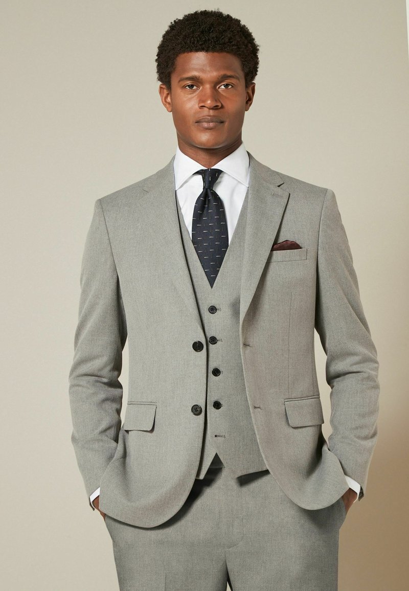 Traje gris de tres piezas con solapas muesca, chaleco abotonado y corbata de patrón oscuro. El pañuelo de bolsillo añade un detalle en contraste. Ajuste suave y entallado.