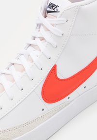 Sneaker alta bianca con parte superiore in pelle liscia, accentuata da un vivace swoosh arancione di Nike e un pannello in suede beige testurizzato sulla suola.
