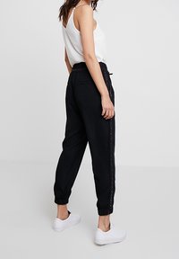 Kobieta w czarnych dresowych spodniach z bocznym paskiem i białym bezrękawnikem, stojąca na gładkim jasnoszarym tle.