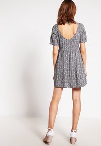 Black patterned dress with short sleeves and a deep back. Flared skirt with a gathered waistline. Paired with white block-heeled sandals.