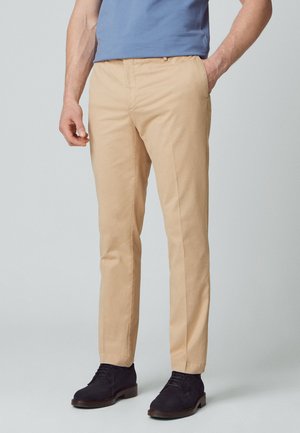 Pantalons beiges à jambes droites avec une texture en coton lisse, poches avant et passants de ceinture, assortis à des chaussures en cuir suédé noir.