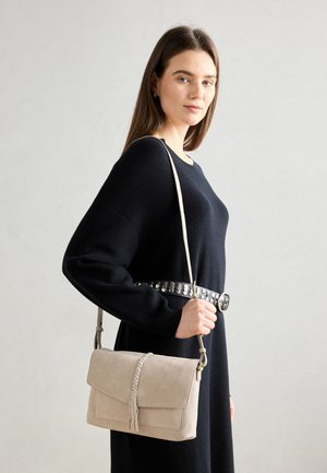 Bežsuede crossbody kott, millel on punutud klapp, reguleeritav rihm ja tähnide detail. Kandmine tumeda sviitri ja studiga vööga.