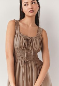 Braunes ärmelloses Kleid mit gerafftem Oberteil, quadratischem Ausschnitt und Binderdetail. Verfügt über gesammelten Stoff und strukturierte Träger.